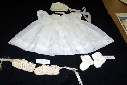 A pair of creamy white knitted wool booties, 1964, with nylon ribbon threaded around ankles. 