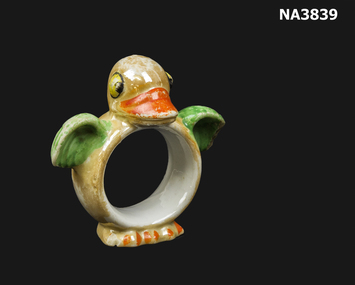Round china ring with duck head & wing on top & feet. Ring is beige, wings green and ducks bill and feet are orange.