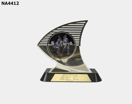Irregular shaped metal trophy on plastic base. Trophy has illustration of two girls playing softball. A metal strip is attached to the base with 'NDSSSA Softball 2nd Blackburn East 1970' inscribed on it.