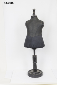 Small child size mannequin on wooden stand. The model is without arms and torso size. It is covered in black cotton and black linen like material above the waist. 