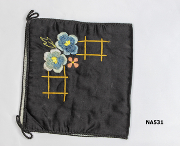 Black sachet with embroidered flowers . Lined with blue material. Bound with black cord looped on two front corners. Probably used to hold small items.