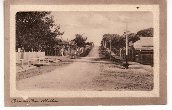 Black and white postcard titled 'Blackburn Road, Blackburn'.  Scene shows Blackburn Road looking  South from railway gates.  Shop on one corner.  Unmade road.