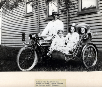 Black and white photo of an adult male with a motorcycle and cane side-car with three children outside Blackburn Hall