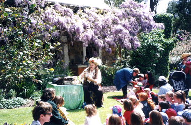 Jason, the Teddy Tale Man and children listening to his story in the garden of Schwerkolt Cottage with Barbara Keene distributing lollies.