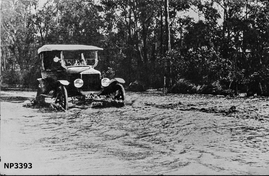 Large black and white photograph of Blackburn Road near Heath Street. A car driving through water with street behind