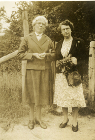 Two women standing in front of a wire fence with trees in the background. 