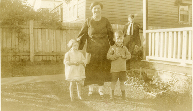 One woman in dark dress standing between two children; on her left a small boy wearing a jersey and short trousers.