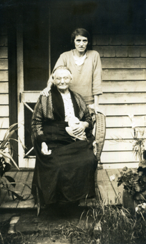  Woman standing behind an older woman who is sitting in a cane chair on a  wooden verandah.