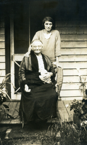  Woman standing behind an older woman who is sitting in a cane chair on a  wooden verandah.
