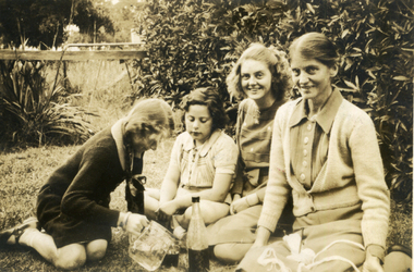 Three woman and a female child seated on the ground in front of a hedge.