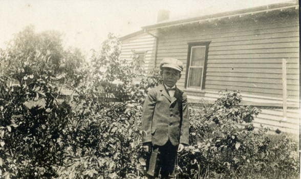 Alwyn Till wearing a cap and standing in the garden in front of a weatherboard house.