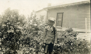 Alwyn Till wearing a cap and standing in the garden in front of a weatherboard house.