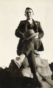 Sydney Till wearing a suit and holding a hat while sitting on a rock.