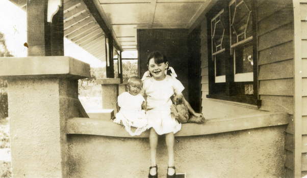 Young dark haired girl in a white dress sitting with a doll and teddy bear on the wall of a verandah.