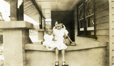 Young dark haired girl in a white dress sitting with a doll and teddy bear on the wall of a verandah.