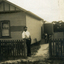Man standing at opening in picket fence on front of wooden house with watertank -