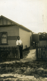 Man standing at opening in picket fence on front of wooden house with watertank -
