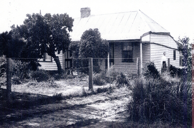 Weatherboard house with a post and wire fence along the drive, and woman standing near the side of the house