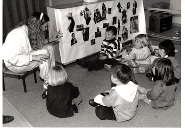 Photograph of a kindergarten teacher playing a game with six small children sitting on the carpet.