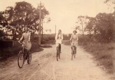 Three girls riding bicycles on an unmade road. One of the girls is Alison Till.
