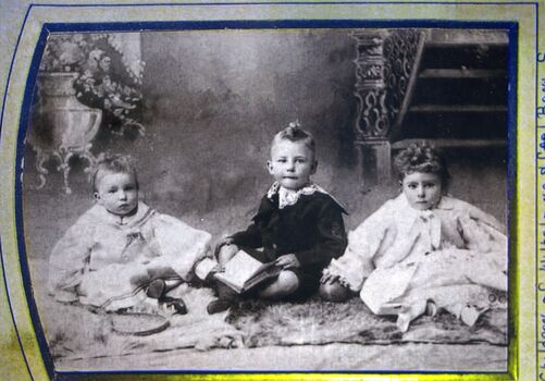 Black and white photograph of two young children in light clothing and a boy in dark sitting on a rug.