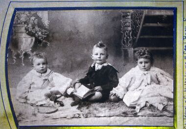 Black and white photograph of two young children in light clothing and a boy in dark sitting on a rug.