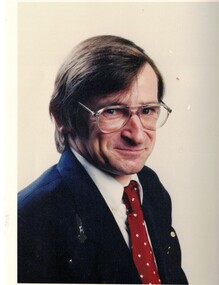 Noel Spurr. Wearing dark suit and burgundy tie with yellow dots. Brown hair and wearing glasses.