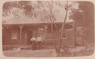Sepia photograph of a wooden house with two people standing in front. 