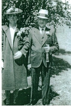 Black & White photograph of a man and woman dressed as if attending a wedding. The woman is carrying flowers and the man has a flower in his buttonhole.