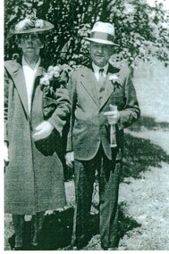 Black & White photograph of a man and woman dressed as if attending a wedding. The woman is carrying flowers and the man has a flower in his buttonhole.
