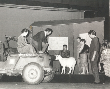 Stage with several actors sitting on a car and standing around and looking at a goat in the centre.