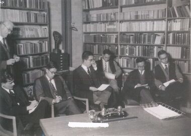 Five seated male students dressed in suits and academic gowns with an older man, also dressed in a suit and academic gown.