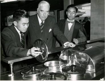 Two male residents of International House wearing suits and academic gowns looking at kitchen equipment with the Prime Minister of Australia Robert Menzies.