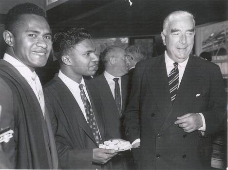 Two male students dressed in suits and academic gowns smiling and standing beside the Prime Minister of Australia dressed in a double-breasted suit