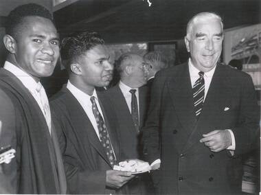 Two male students dressed in suits and academic gowns smiling and standing beside the Prime Minister of Australia dressed in a double-breasted suit
