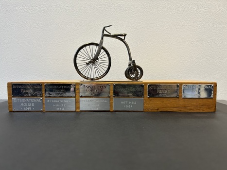 Miniature metal tricycle mounted on a timber base, metal plaques around edge of base with winners of the race (different university colleges and departments) from 1965 to 1983.
