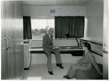 Two men in suits sit in a room and talk, one on the desk at the window, and one on a mattress on the floor.