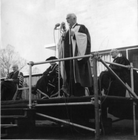 Black and white photograph of a white-haired man dressed in a suit and an academic gown standing on a podium. 