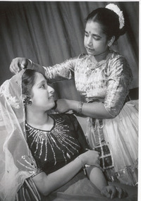 Two female students from Malaysia dressed in traditional costume. One is standing and adjusting the veil of the other, who is sitting.