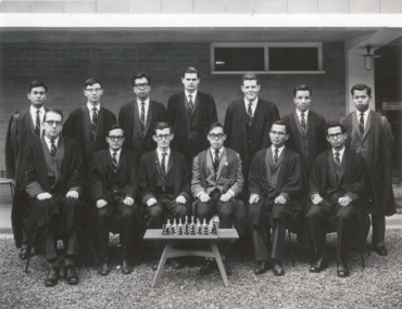 Thirteen men pose in suits and academic gowns, six sitting and seven standing behind a chess board