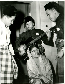 Three male students, one of whom holds a guitar, stand around a kneeling female student. They are all smiling.