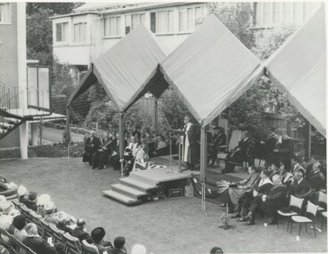 An awning over small stage with a man at a microphone in an Oxford style graduate gown. The crowd sitting in chairs on either side of him are also wearing Oxford style graduate gowns. He is speaking to a seated audience in front of him.  