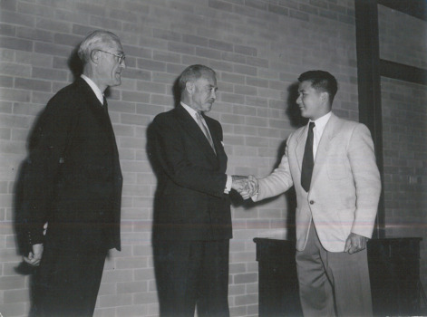 Two men shake hands while another man watches to the left