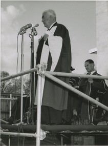 A man in his sixties in a traditional Oxford graduation gown stands on a podium speaking into a microphone. A few people in graduation gowns stand behind him. 