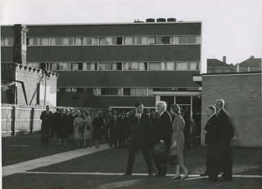 A man in a suit leads another man and his wife down a path on the International House campus. Two more suited men trail behind them. In the distance there is crowd of well-dressed men and women. The are in the courtyard between buildings. 