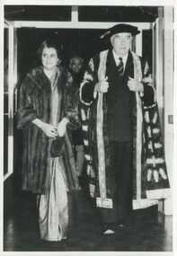 An older male (Chancellor Robert Menzie) in an Oxford graduate robe walks through a entrance of a building with a women in a fur coat (Prime Minister of India Indira Gandhi) 