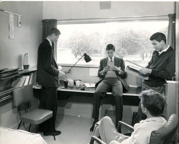 Four men sit and stand in a circle in front of a student room desk, a window looks out onto Royal Parade