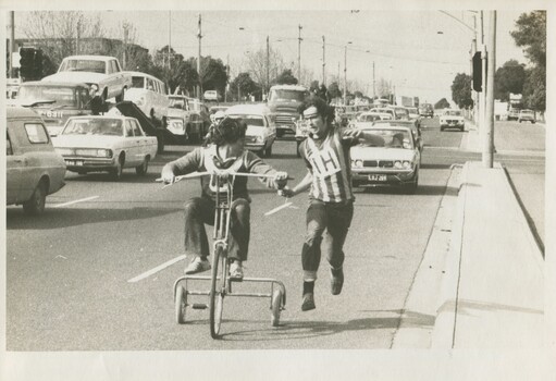 A young man riding a poorly made tricycle next to another young man running in a striped sports jersey. The running young man looks concerned at the bike's stability. They are on a main road with lots of cars. 