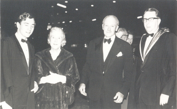 Four people (three men and one woman) stand in a row looking at the camera. They wear formal dress.