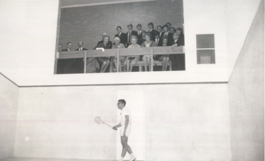 Man plays squash in a white court while a group of formally-dressed men and women watch from seating above.